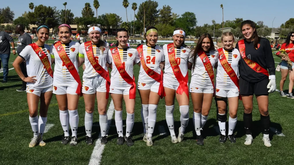 Girls soccer team posing on the field, wearing white uniforms with red sashes that read 'Senior' celebration.