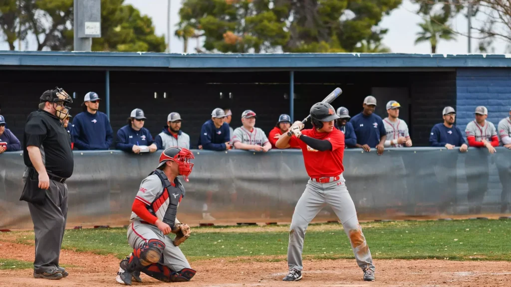 Baseball batter in red swings at a pitch as catcher crouches behind home plate and umpire watches nearby.