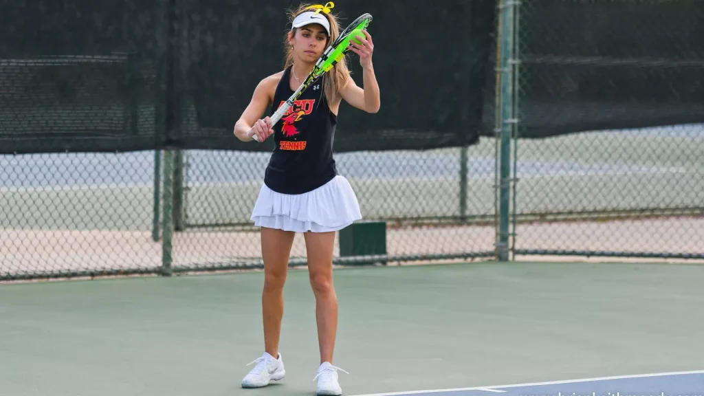 Female tennis player on a court, preparing to hit a shot with a neon-green racket, wearing a black sleeveless top, white skirt, white shoes, and a white visor with a yellow bow.