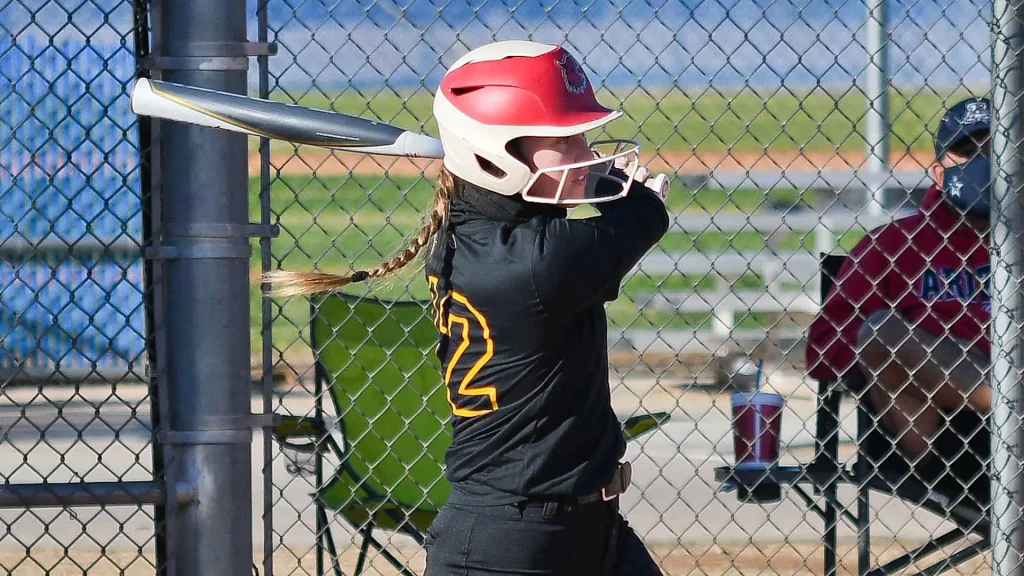 A girl in a black and orange baseball uniform winds up to bat, wearing a red-and-white helmet, with braided hair visible beside a chain-link fence.