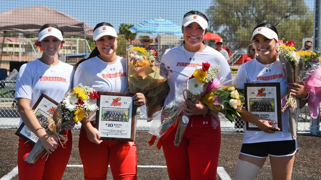 Four Firestorm softball players hold bouquets and framed awards on a sunny field.