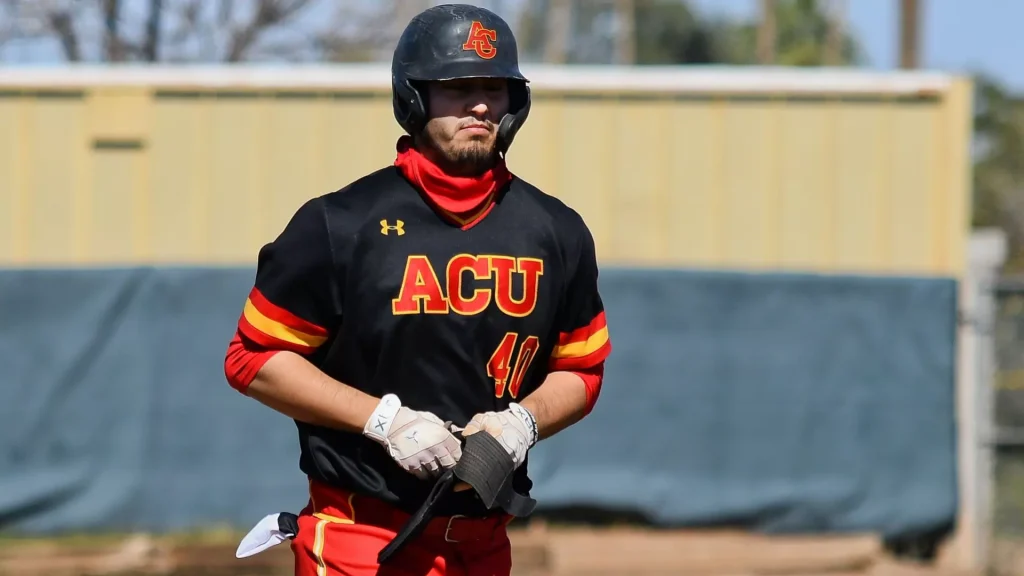 Baseball catcher in a black and red ACU uniform with helmet, standing on the field with gloves on.