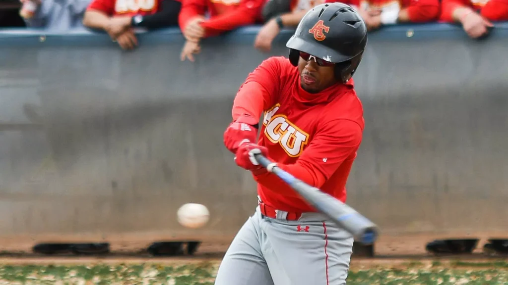 Baseball player in a red uniform swings at a pitch, wearing a helmet, with fans in the background.