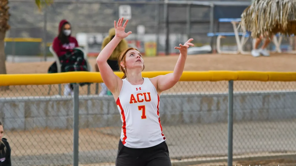 Female athlete in a white ACU jersey (number 7) reaching upward during a play near a yellow padded fence behind a chain-link fence.