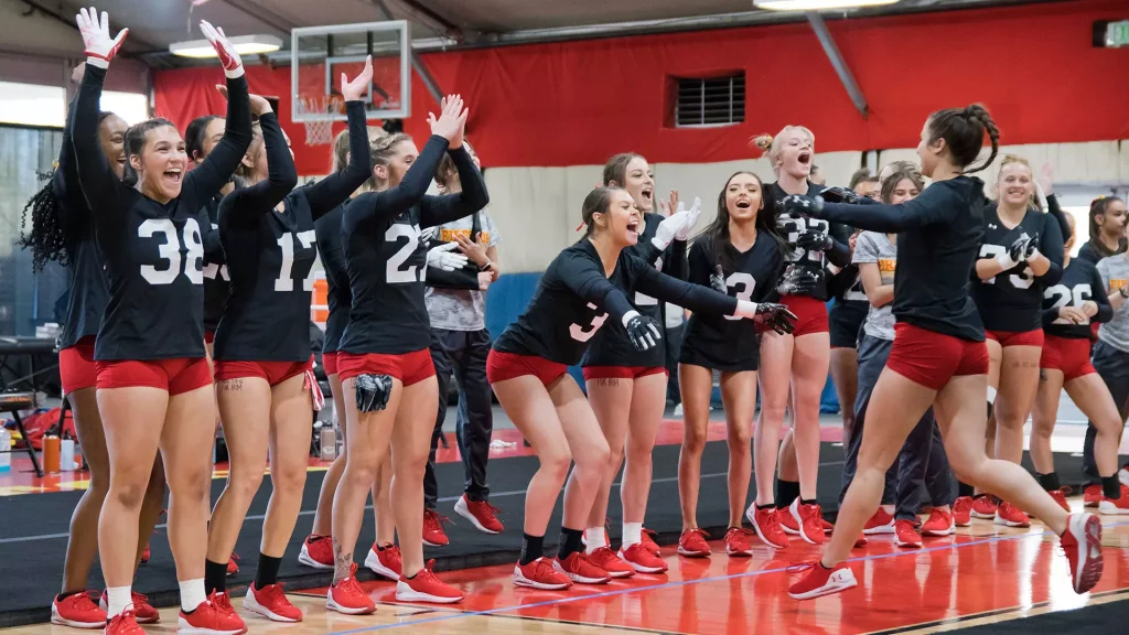 A large group of female volleyball players in black jerseys and red shorts celebrate, raising hands and cheering on a gym floor.