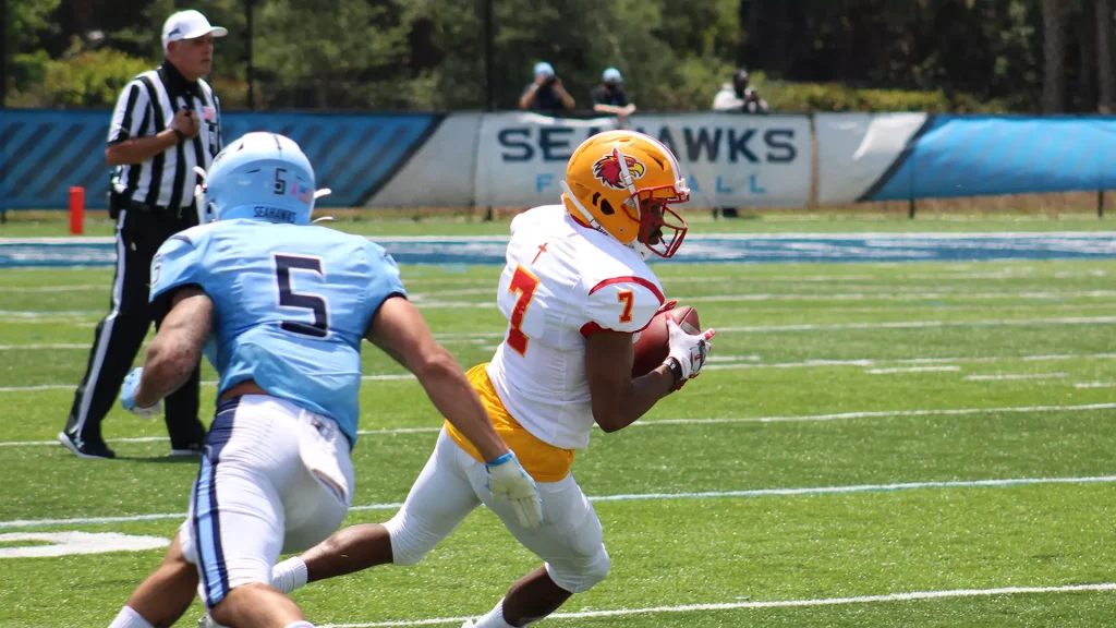 American football player in white and gold uniform runs with the ball while a defender in light blue tries to tackle him on a sunny field in action.