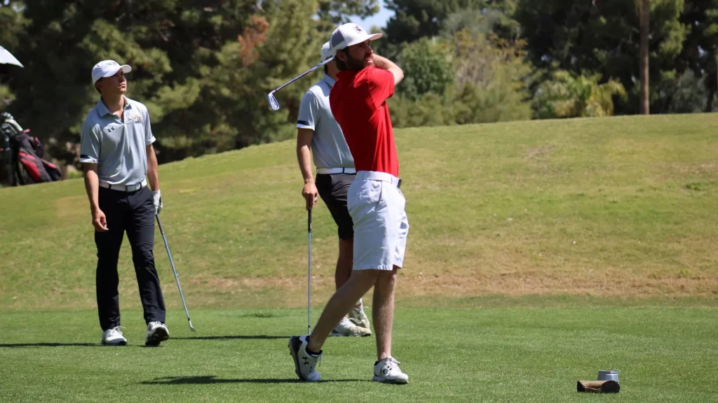 Golfer in red shirt mid-swing on a sunny golf course, with two players watching nearby.