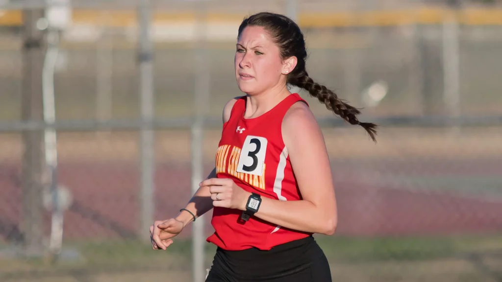 Female runner in red athletic top with bib number 3, mid-stride on a track field.