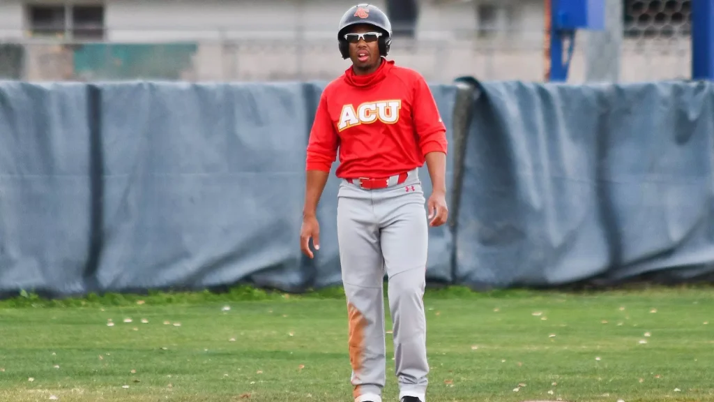 Baseball player in a red ACU jersey stands on a grass field with a padded barrier in the background.