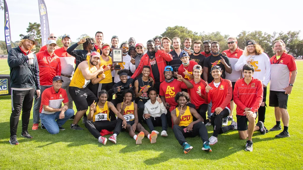 Group of diverse athletes and coaches on a sunny sports field posing with a championship plaque at center, wearing red and yellow team uniforms.