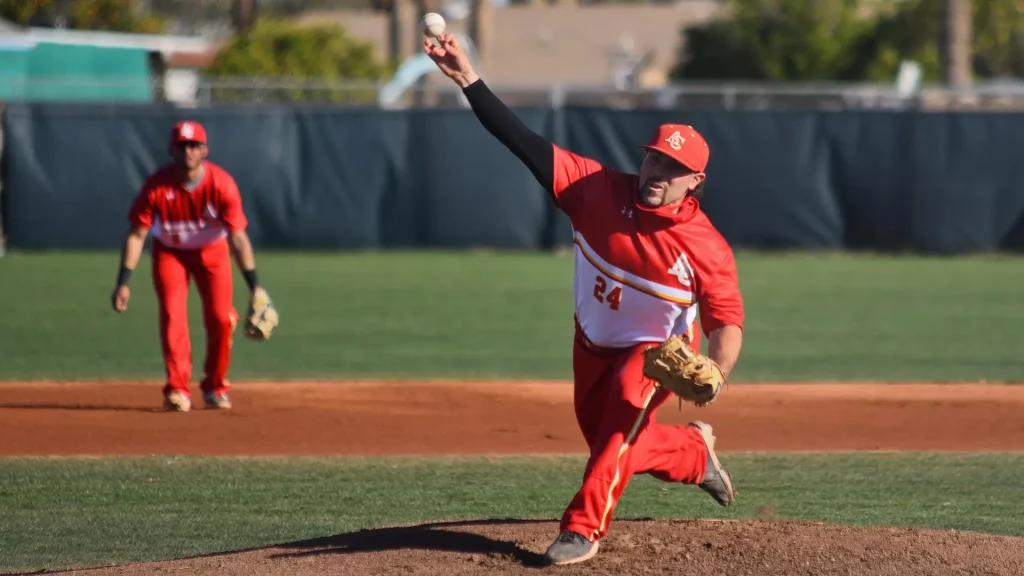 Baseball pitcher in red and white uniform winds up and throws a pitch on the mound, with a fielder in red in the background.