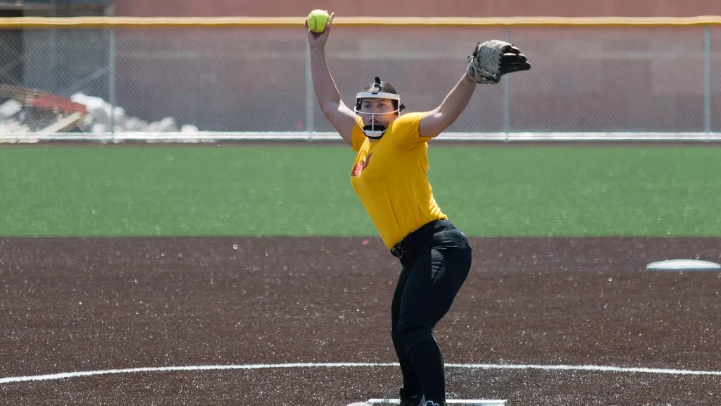 Softball pitcher in a yellow jersey mid-throw, right arm raised with a green ball, on a dirt field.