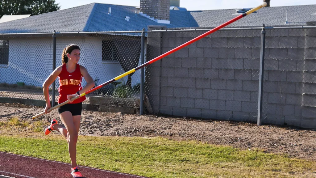 Female athlete in red top and black shorts runs on a dirt track while holding a javelin, outdoors by a chain-link fence and buildings behind her.