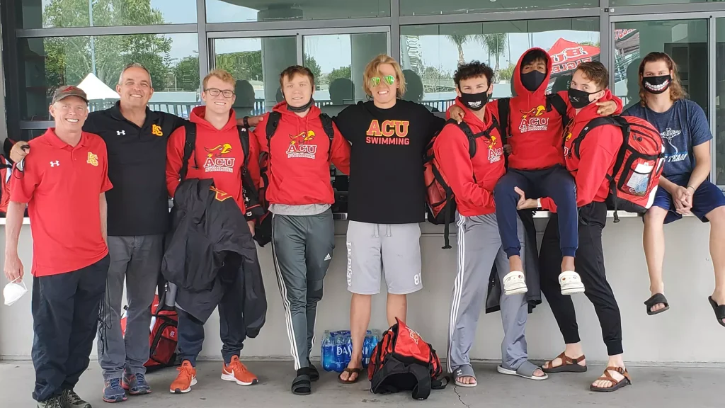 Group of athletes in red ACU swim shirts posing together outside a building, smiling at the camera with backpacks and water bottles nearby.