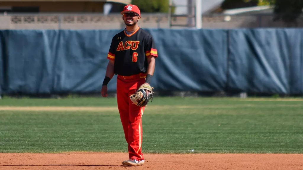 Baseball player in black ACU jersey (number 6) and red pants stands on the infield with a glove in hand.