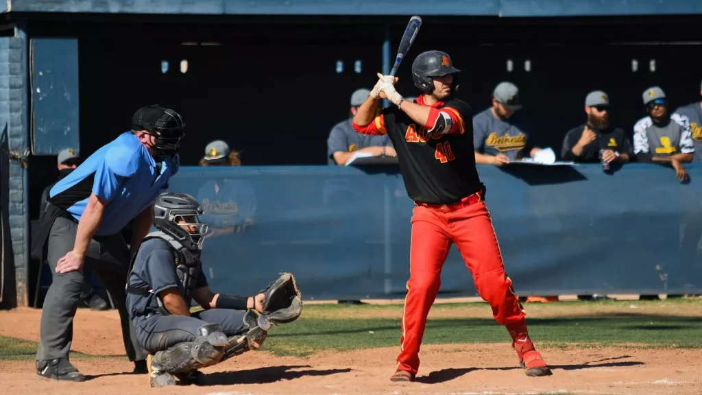 Baseball batter in black and red swings at a pitch as the catcher crouches and the umpire watches from the left on a dirt infield.