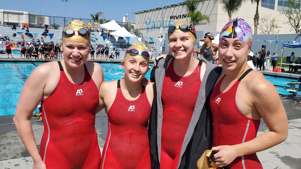 Four young female swimmers in red suits posing together by a pool after a race, wearing swim caps and goggles on their foreheads with a cheering crowd in the background.