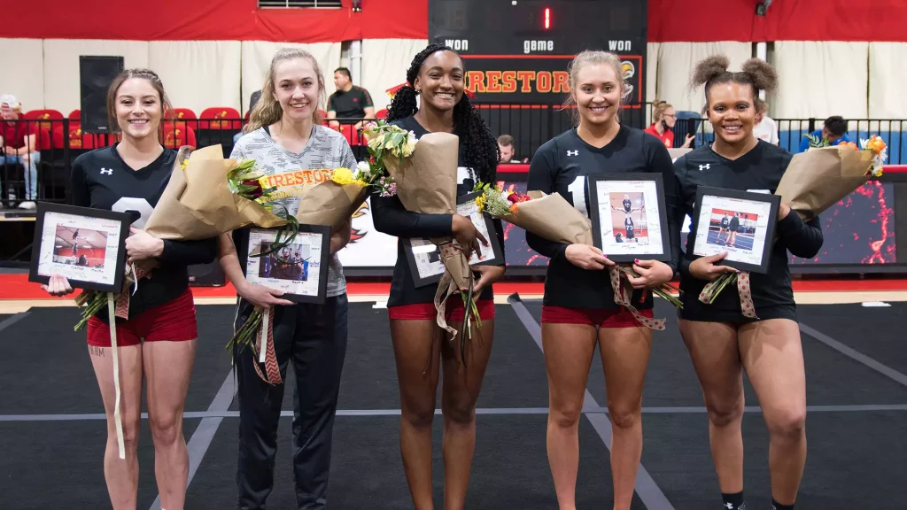 Five young female athletes stand together on a gym floor, each holding a bouquet and a framed award photo, smiling for the camera in an awards moment.