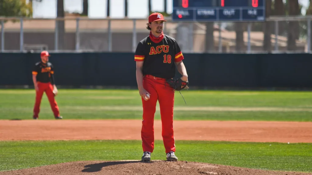 Baseball pitcher in a black and red ACU uniform stands on the mound with a ball in his right hand and a glove on his left hand.
