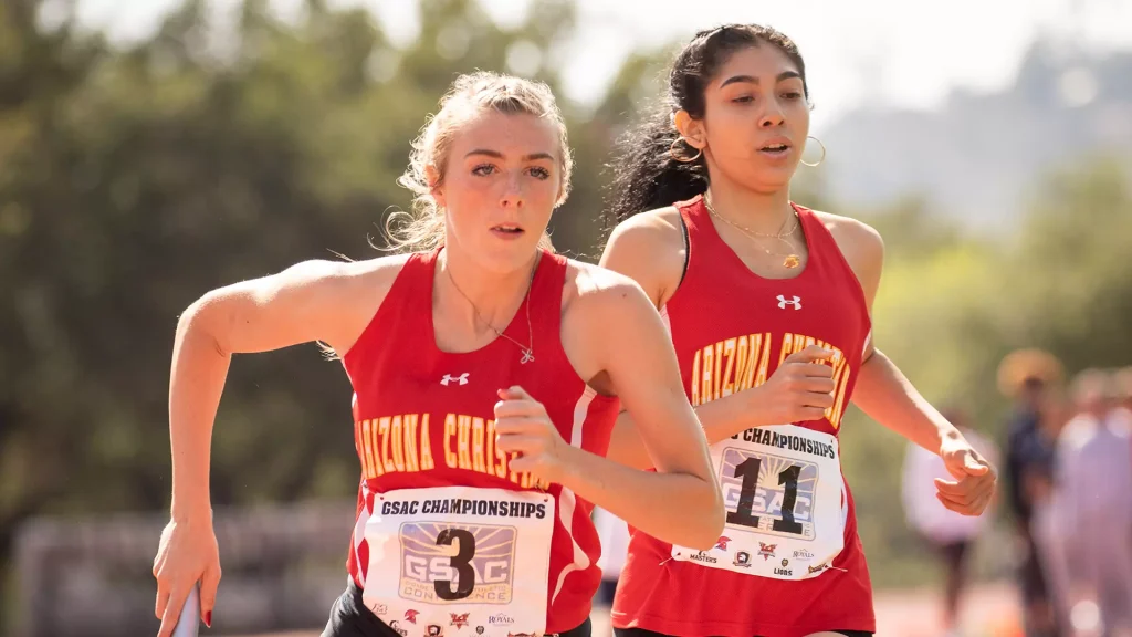 Two female runners in red uniforms sprint side by side during a race, bib numbers visible on their chests.