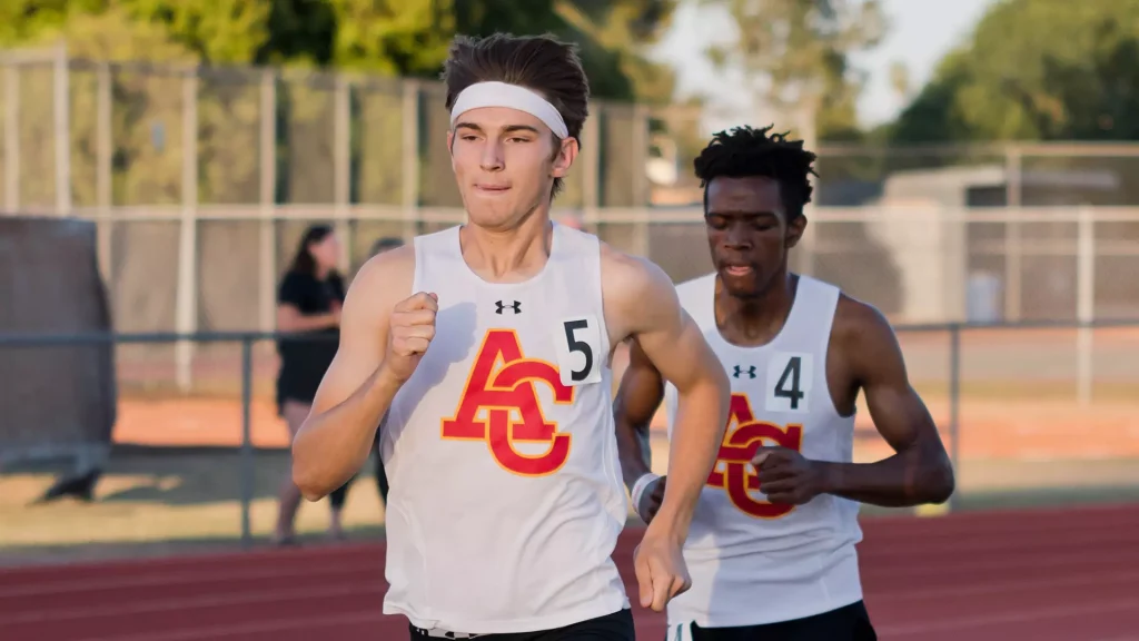 Two male runners in white singlets with red and gold school logo sprint on a track, wearing numbers 5 and 4.