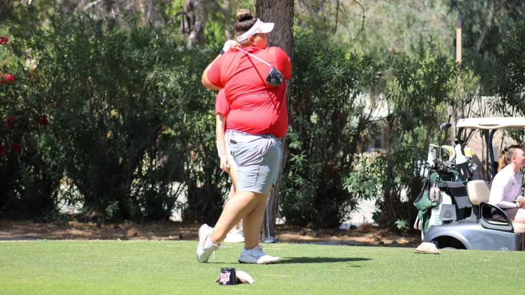Golfer in a red shirt and white visor walks on a tee, carrying a club over her shoulder; golf cart with bags visible in the background.