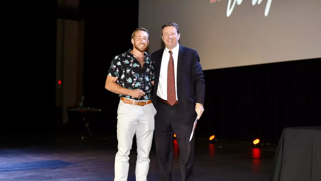 Two men on a stage posing together for a photo, one in a tropical shirt and light pants, the other in a dark suit with a red patterned tie.