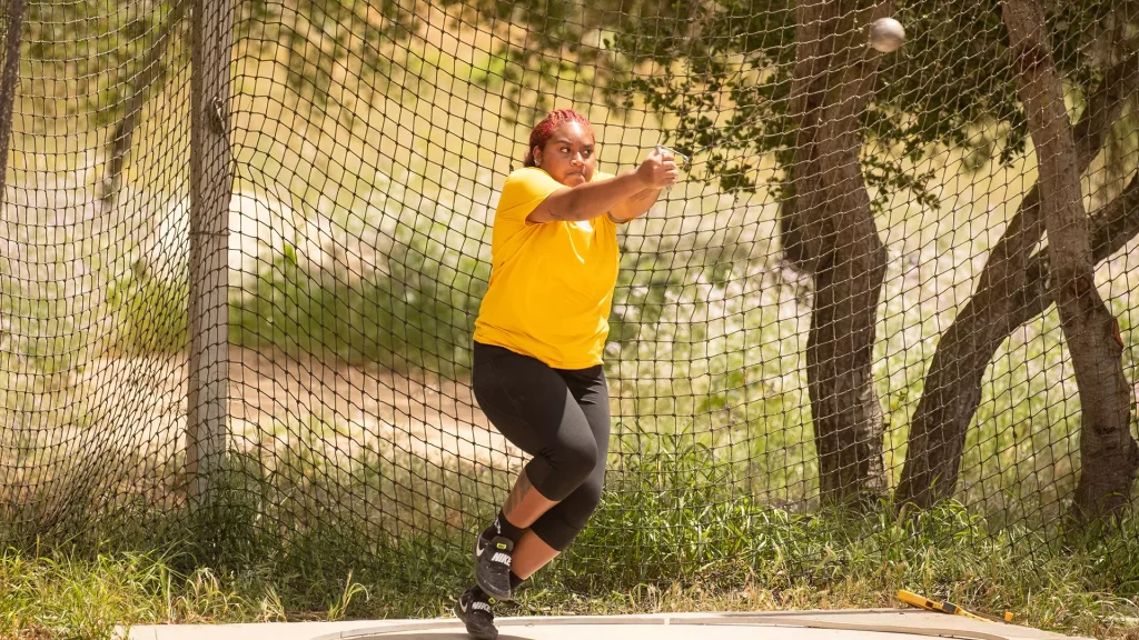 Athlete in a yellow shirt and black leggings throws a ball inside a chain-link fenced batting cage outdoors.