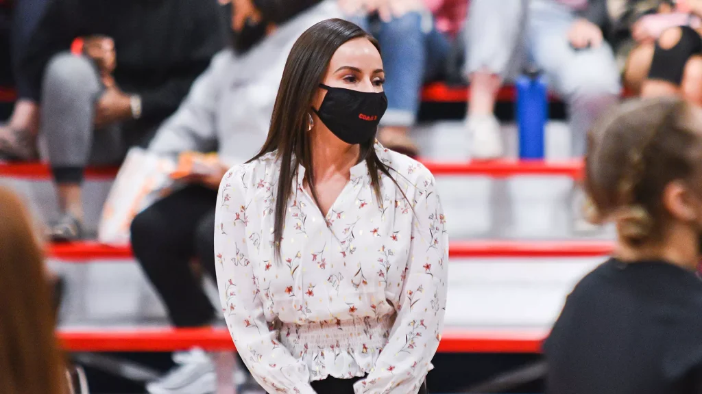Woman wearing a black face mask and white floral blouse in a crowded public space at an event, facing to the side.