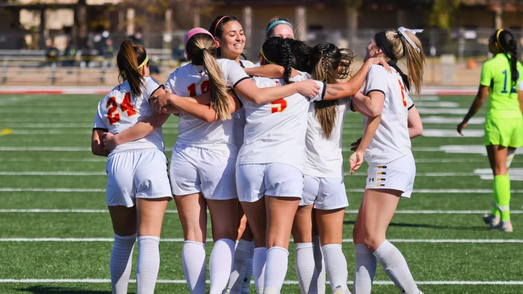 Female soccer players in white uniforms huddle in a circle on a green field, arms around each other, celebrating together.
