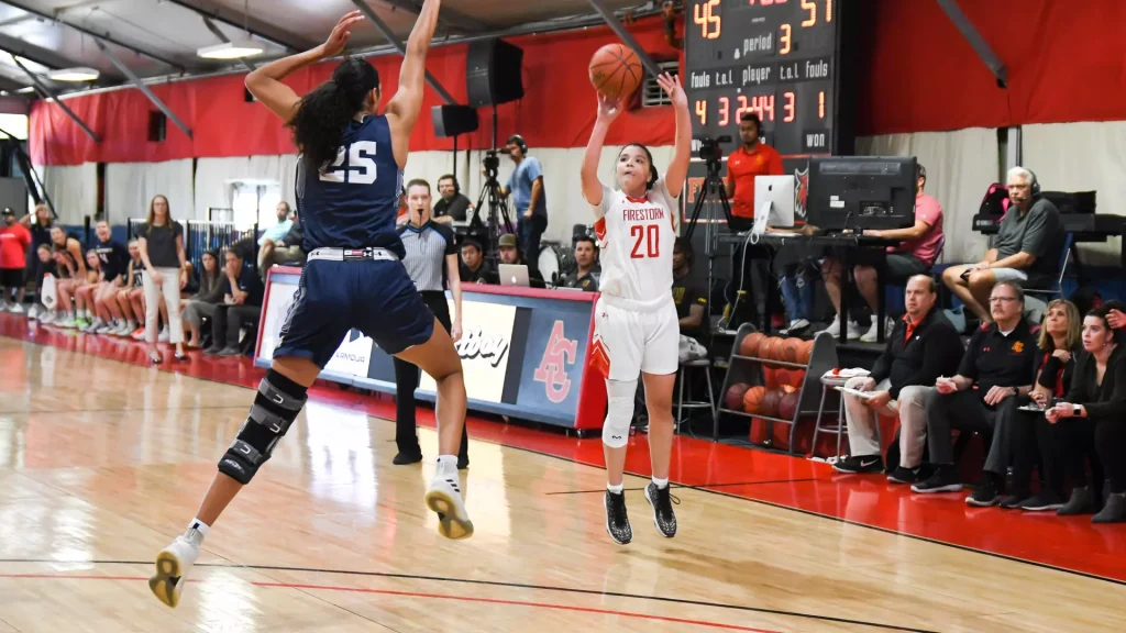 Girls' basketball game in a gym; player in white jersey number 20 jumps to shoot or pass while defender in dark blue jersey number 25 leaps to block, near the basket.