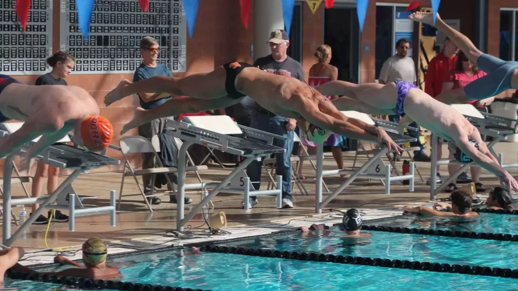 Swimmers dive from starting blocks into a pool during a race, with spectators watching in the background.