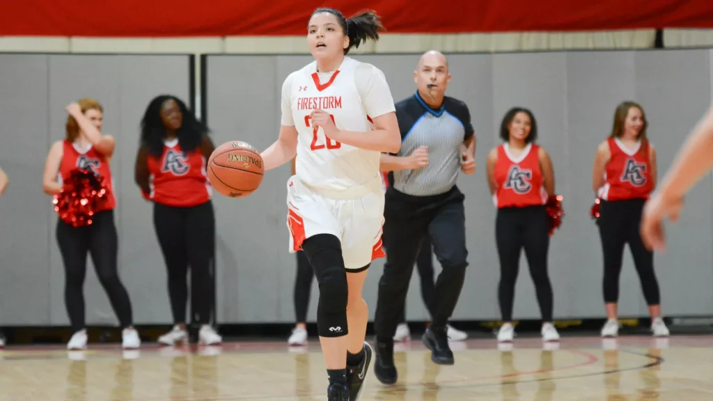 Female basketball player in a white Firestorm jersey (number 20) dribbles down the court as a coach and cheerleaders watch in the background.