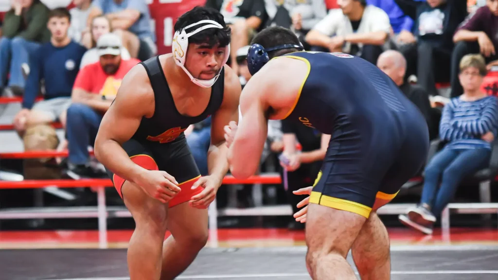 Two male wrestlers grapple on a red-matted mat as spectators watch from the stands, one in a black singlet with red trim and headgear, the other in blue with yellow trim.