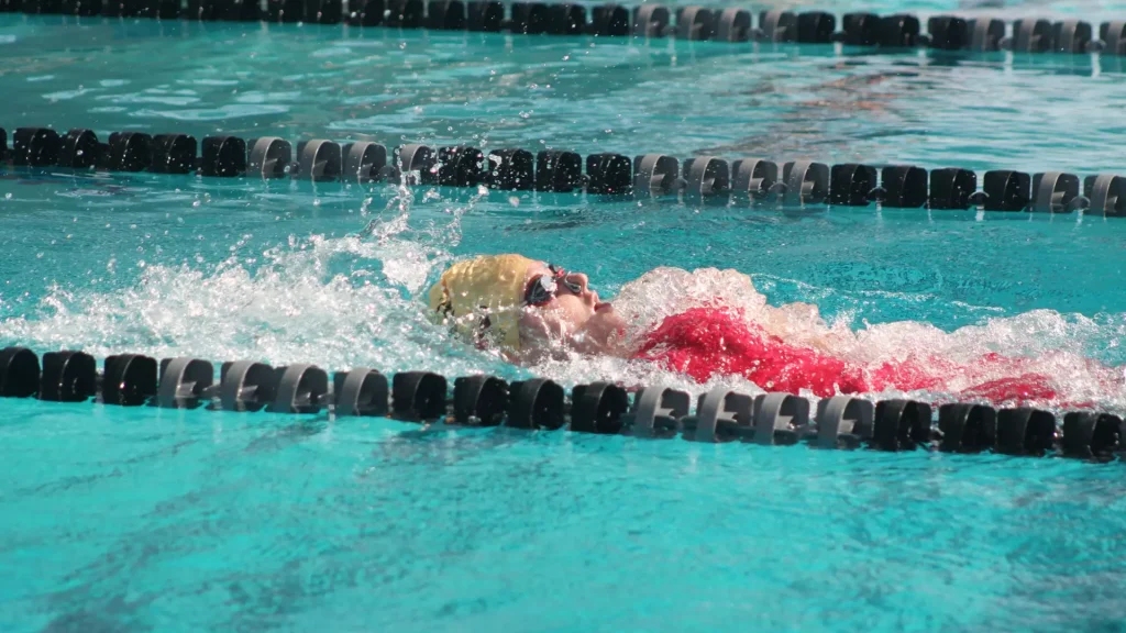 Firestorm Record More National Qualifying Times In Battle At Grand Canyon - Arizona Christian University Swimmer mid-stroke in a turquoise pool wearing a yellow cap and red suit, with lane dividers nearby.