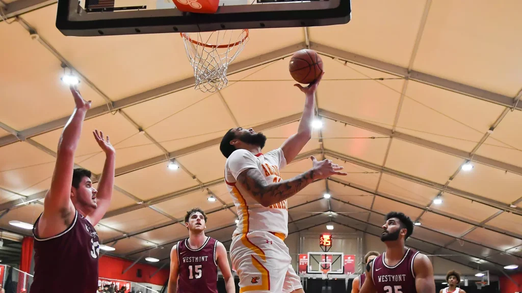 Basketball player in white and orange drives to the basket for a layup as defenders in maroon Westmont jerseys contest the shot in a bright gymnasium.