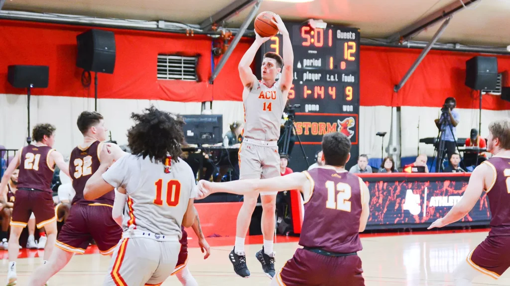 Player in gray jersey (#14) jumps to shoot while defenders in maroon surround him in a gym‑style basketball game. Scoreboard in background shows time 5:01 and ACU lettering on jersey.