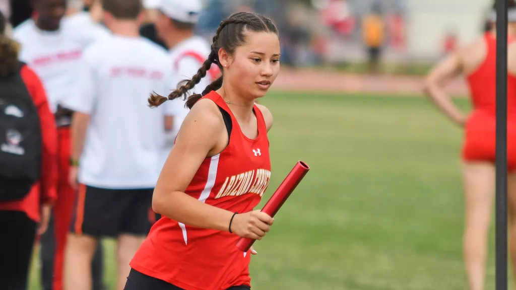 Female athlete in a red uniform running with a relay baton during a track relay race on the field.