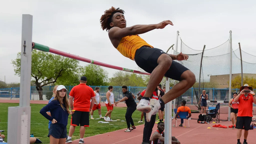 Male high jumper in an orange top clears a red bar during a track meet, with spectators and coaches nearby.