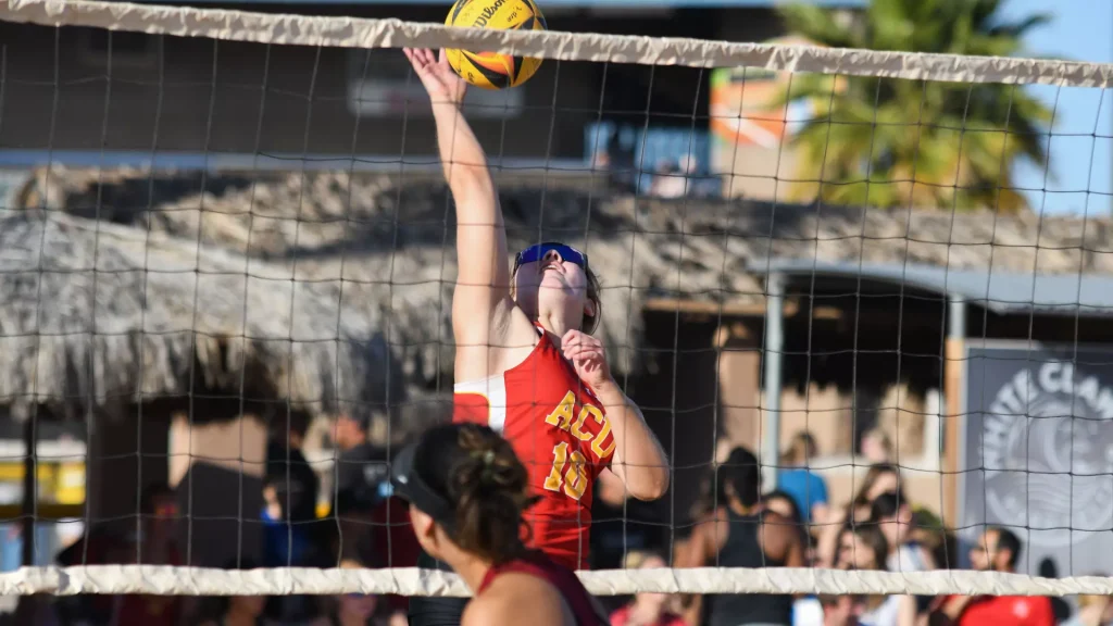 Female volleyball player in a red uniform jumps to spike the ball over the net at an outdoor match, with spectators in the background.