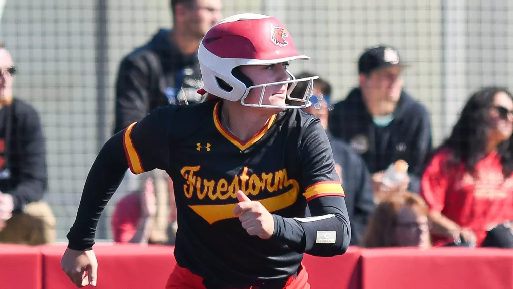 Softball batter in a black and gold 'Firestorm' uniform with a red helmet, mid-swing during a sunny game. Behind her, spectators watch through a fence.