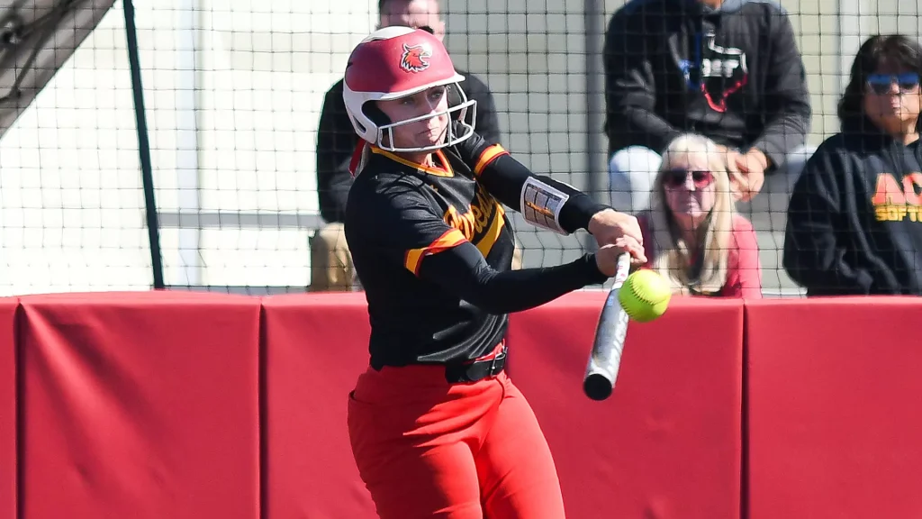 Female softball player in black and orange uniform swing-hitting a bright yellow ball, helmet with faceguard, behind a protective red barrier with spectators in the background