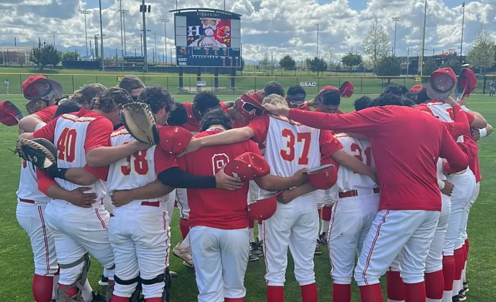 Baseball team in red and white uniforms huddles together on the field with arms around each other, scoreboard visible in the background.