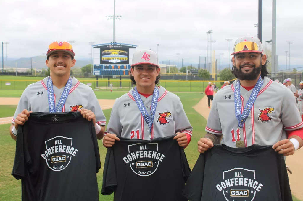 Three smiling baseball players wear All-Conference medals and hold t‑shirts on a field with a scoreboard in the background.