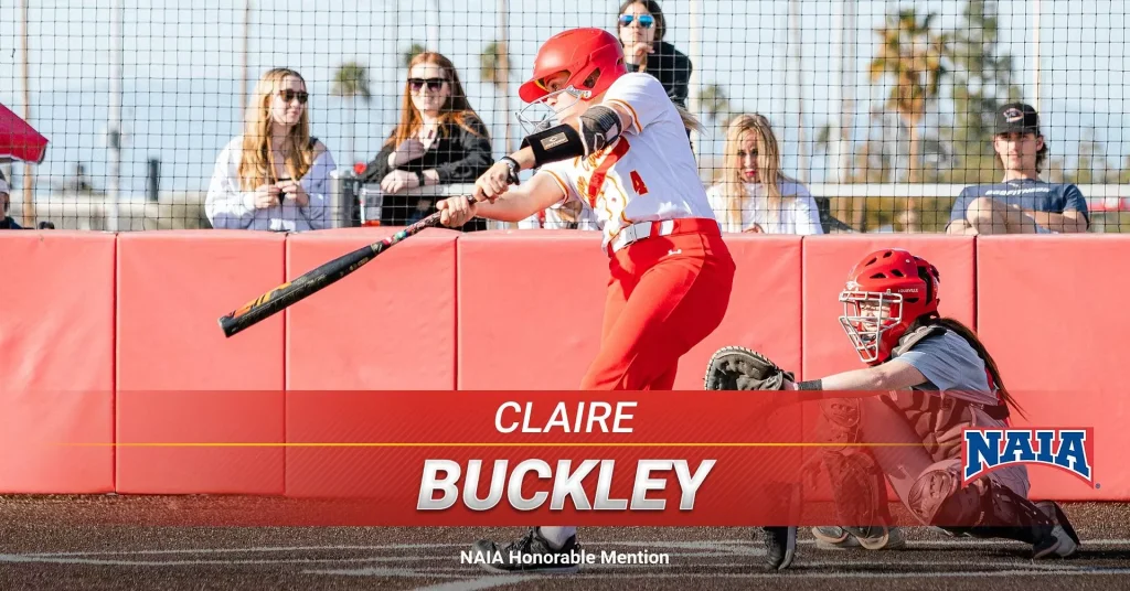 Softball batter in red and white swings at a pitch as the catcher crouches behind a red wall; crowd watches from behind a fence. Banner reads 'CLAIRE BUCKLEY' and the NAIA logo.