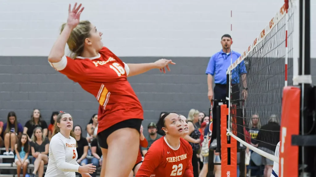 Female volleyball player in a red jersey leaps to spike the ball at the net as teammates and a crowd watch from the stands.