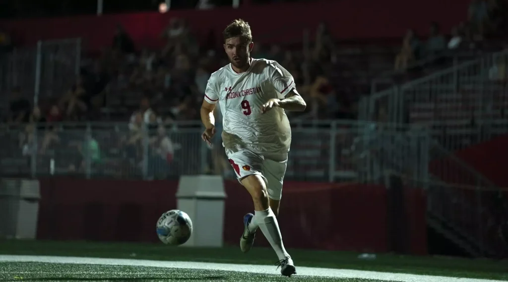Soccer player in a white Arizona Christian uniform #9 dribbling the ball on a night match, stadium lights on.
