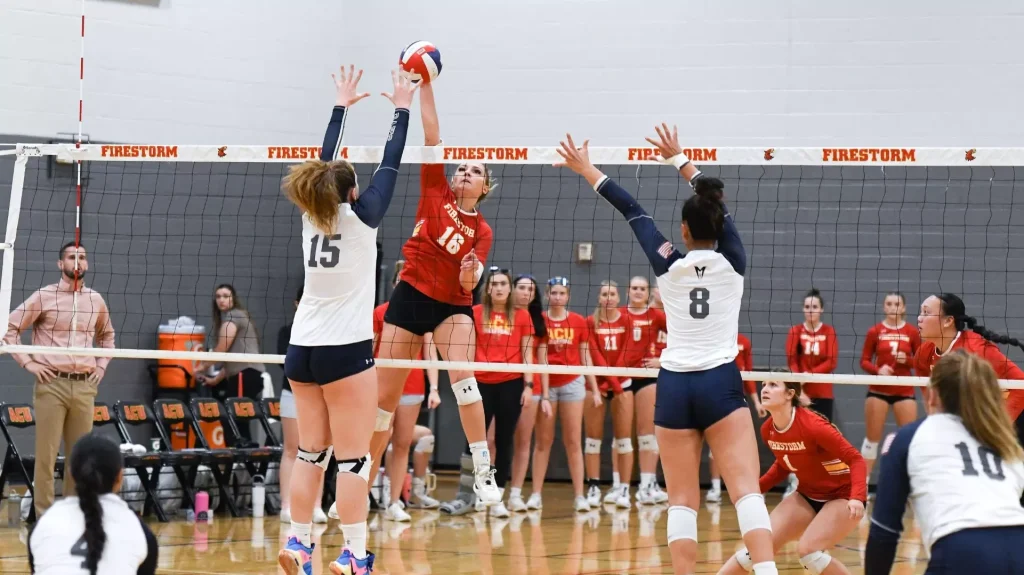 Red-team attacker spikes the ball while two white-team blockers jump to stop it at the net in a gym match.