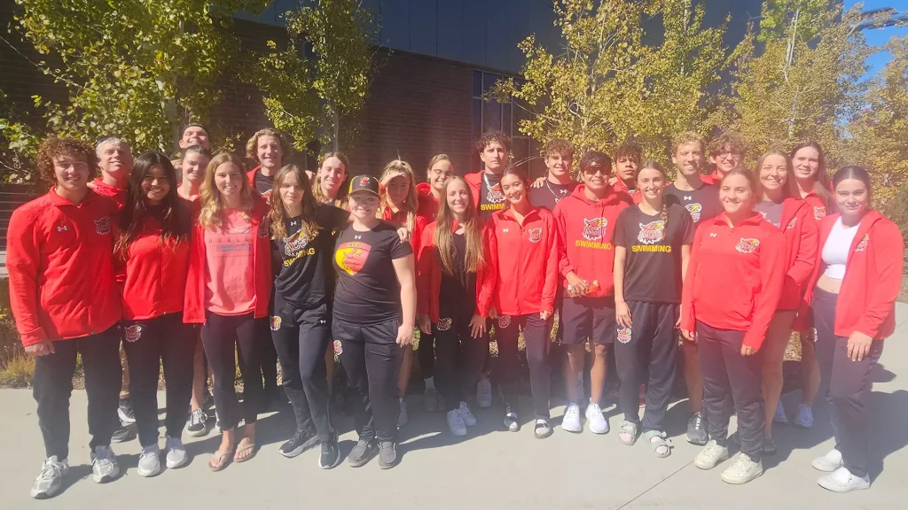 Large group of students in red and black athletic outfits posing outside a brick school building on a sunny day.