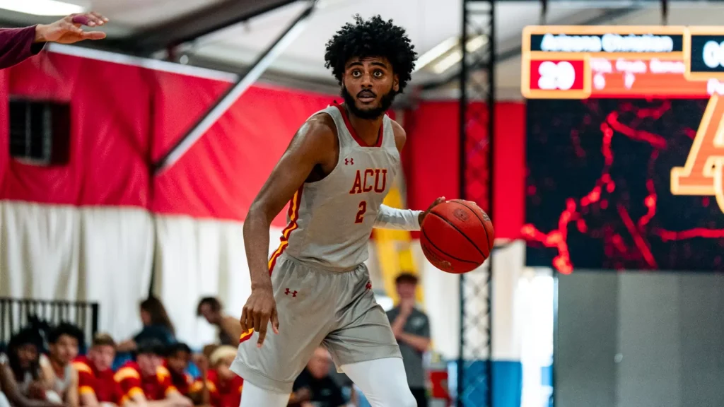Male basketball player in a gray ACU uniform dribbles the ball during a game, crowd in red and gold behind him.